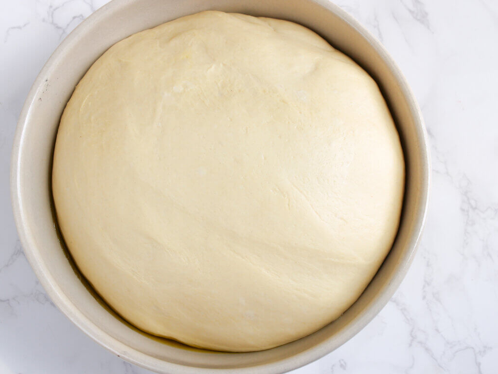 Homemade hamburger bun dough after the first rise, doubled in size and resting in a lightly oiled bowl before being shaped into burger buns.