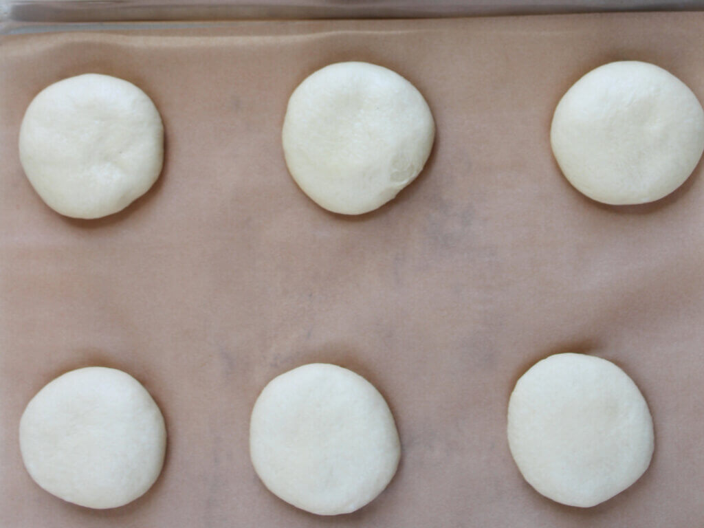 Shaped hamburger bun dough gently flattened by hand and spaced evenly on a parchment-lined baking sheet before the second rise.
