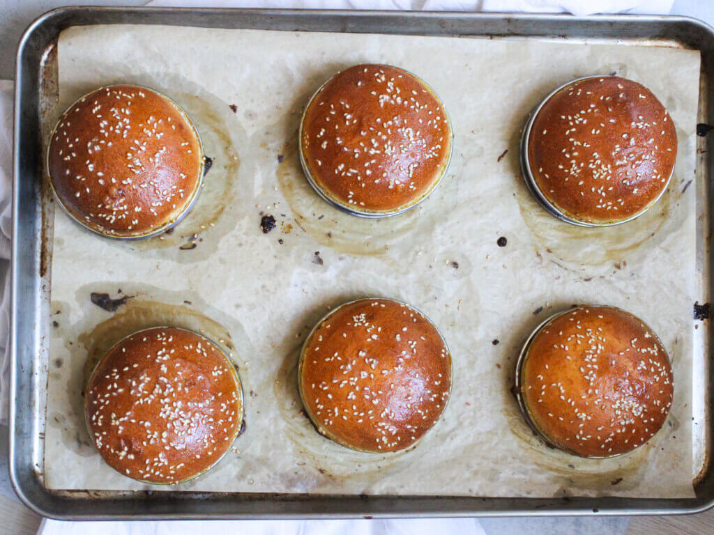 Freshly baked homemade hamburger buns with sesame seeds cooling on a baking sheet after being brushed with butter for a soft, shiny finish.