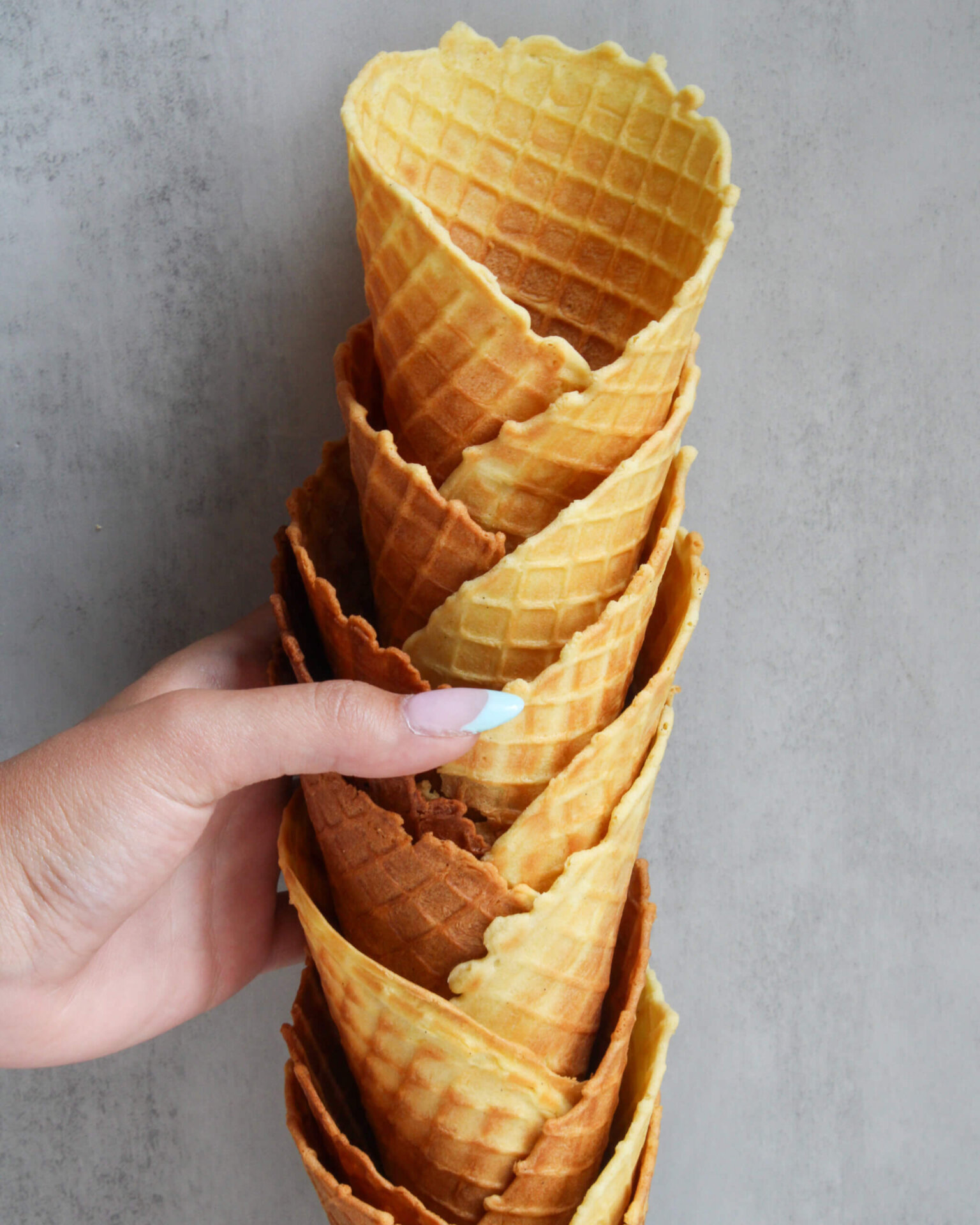 Hand holding a stack of crispy homemade waffle cones with a beautiful golden-brown color and classic waffle pattern against a neutral background. These buttery homemade ice cream waffle cones are thin, crisp, and perfect for filling with your favorite ice cream or frozen desserts.