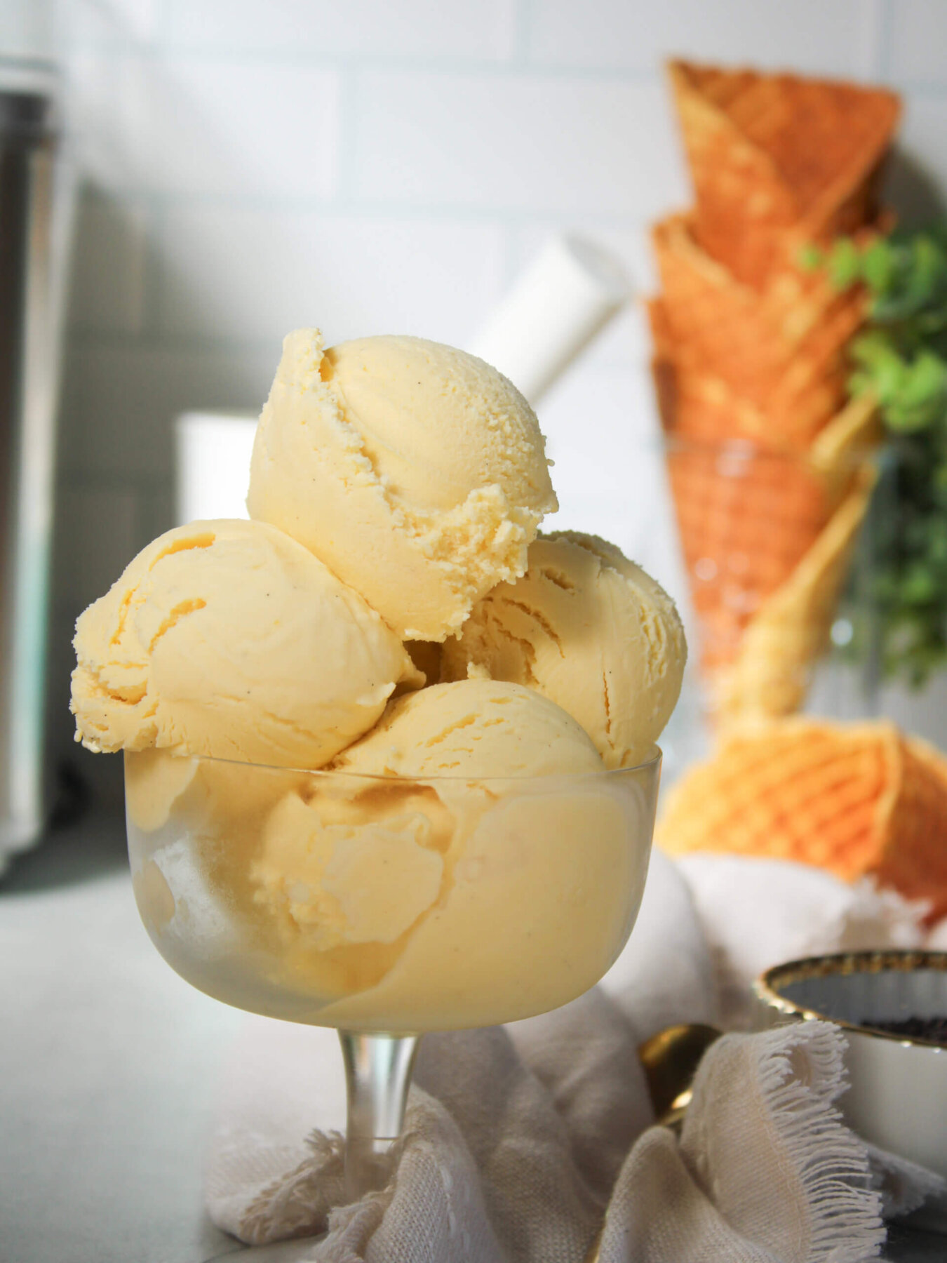 Creamy homemade vanilla ice cream scooped into a clear dessert glass, showcasing its smooth, silky texture and rich vanilla bean specks. Crisp homemade waffle cones in the background add a cozy, old-fashioned touch to this classic homemade vanilla ice cream recipe.