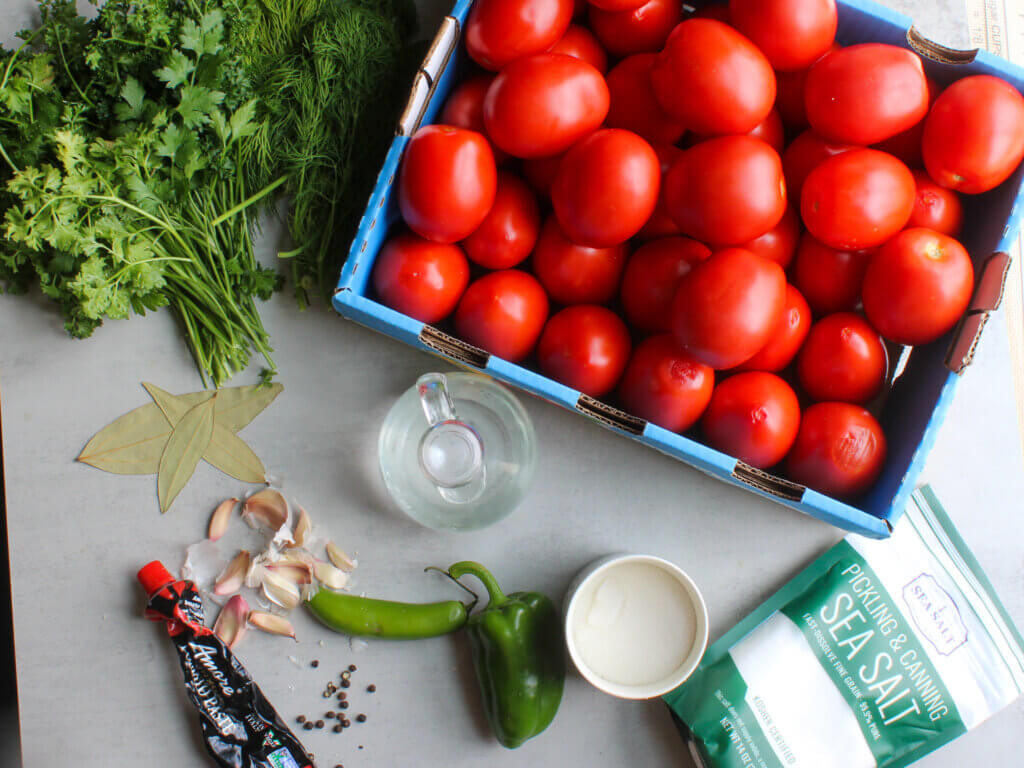 Fresh Roma tomatoes, dill, parsley, garlic, jalapeño, bell pepper, pickling salt, vinegar, and tomato paste for making homemade Russian pickled tomatoes.