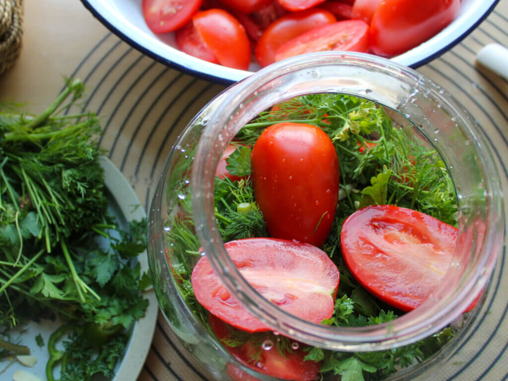 Layering fresh Roma tomatoes with dill, parsley, garlic, and peppers into a glass jar before pouring the pickling brine.